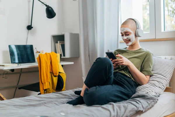 bald female with cosmetic face mask using mobile phone in her room