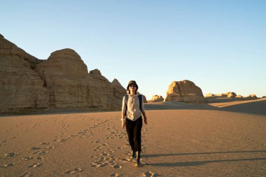 Asyalı kadın gezgin gün batımında çölde yürüyen sırt çantalı ve arka planda yardang bir arazi var.