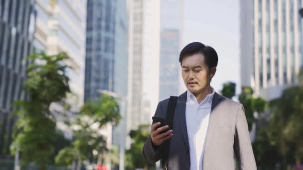 現代都市の携帯電話を見て中央ビジネス街を歩く若いアジア人ビジネスマン — ストック動画