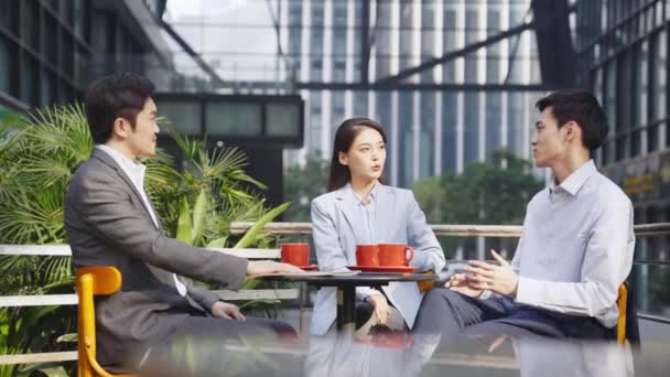 アジア系のビジネスマン2人と屋外のコーヒーショップに座ってディスカッションをしている女性 — ストック動画