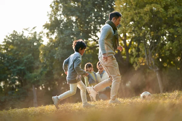 亚洲人的父亲和儿子在公园外面踢足球 而母亲和女儿在后面观看 — 图库照片