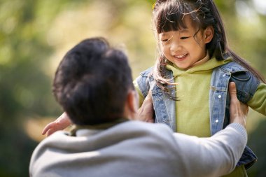 Genç Asyalı baba, 4 yaşındaki kızını parkta kucaklıyor. Mutlu ve gülümsüyor.