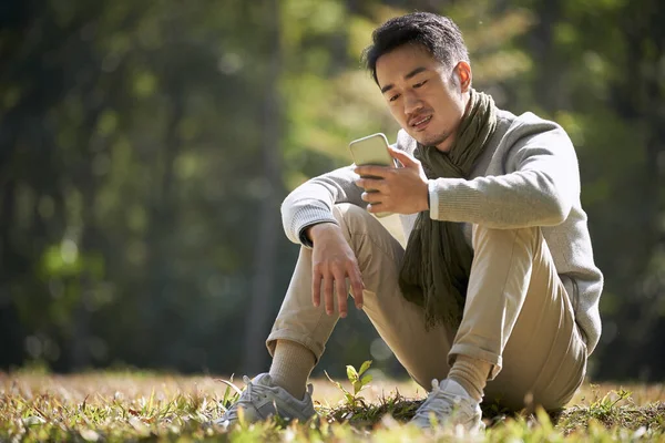 都会の公園で屋外の携帯電話を見て草の上に座っている若いアジア人男性は — ストック写真