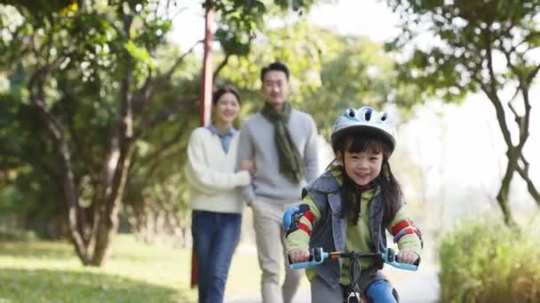 両親が後ろから見ている間公園の屋外で完全な保護具で自転車に乗る小さなアジアの女の子 — ストック動画