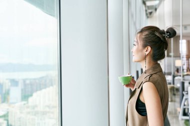 Olgun Asyalı kadın pencerenin kenarında durmuş elinde kahve ya da çay bardağıyla pencereye bakıyor.