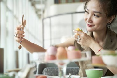 Yetişkin Asyalı kadın restoranda yemek yerken cep telefonu kullanarak selfie çekiyor.