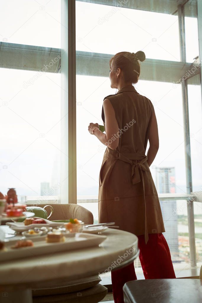mature asian woman looking at view through window of hotel room with breakfast on table