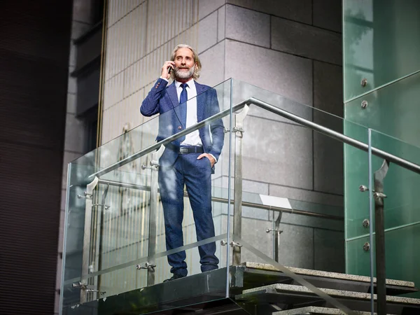 mature caucasian corporate business man standing on top of stairs making a call using cellphone in modern office building
