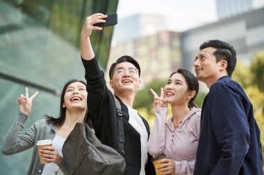 Bir grup Asyalı, cep telefonuyla sokakta mutlu bir şekilde selfie çekiyor ve gülümsüyor.
