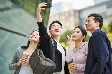 Bir grup Asyalı, cep telefonuyla sokakta mutlu bir şekilde selfie çekiyor ve gülümsüyor.