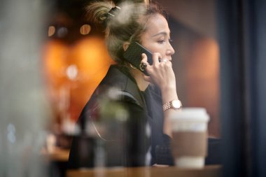 Asyalı bir kadın kahve dükkanında cep telefonuyla sohbet ediyor.