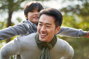young asian father carrying son on back having fun enjoying nature outdoors in park