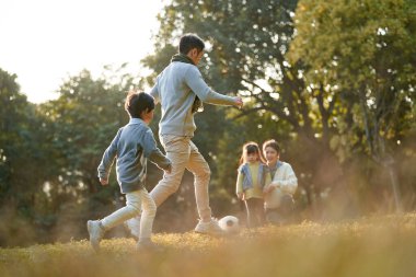 asian father and son playing soccer football outdoors in park while mother and daughter watching on the side
