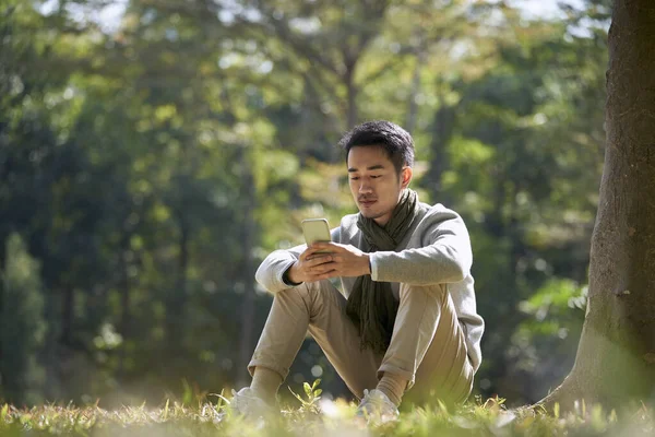 Young Asian Adult Man Sitting Grass Park Looking Cellphone — ストック写真