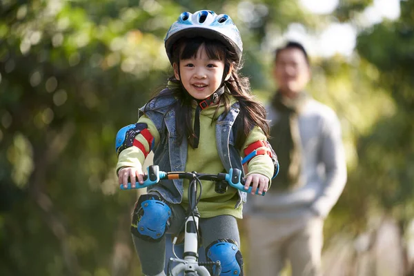 little asian girl with helmet and protection gear riding bike in city park with father watching from behind