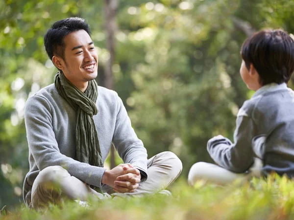 asian father and son sitting on grass having a pleasant conversation outdoors in park