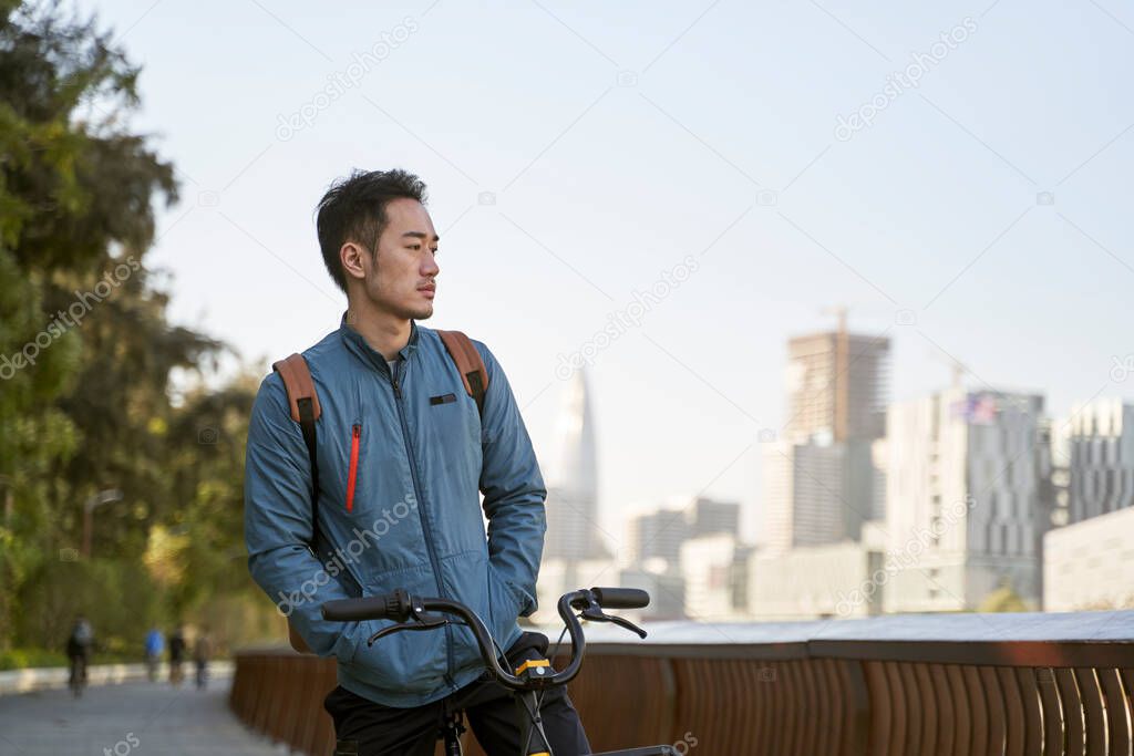 asian young adult man sitting on his bicycle lost in thought in city park