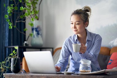 mature professional asian woman working from home sitting in couch drinking coffee while looking at laptop computer