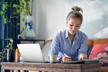 mature professional asian woman working from home taking notes while using laptop computer