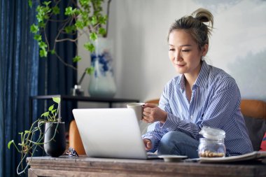 mature professional asian woman working from home sitting in couch drinking coffee while looking at laptop computer