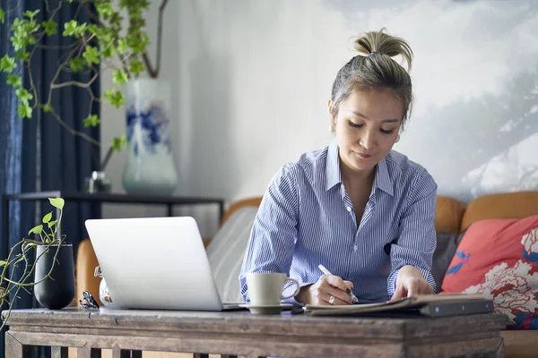 mature professional asian woman working from home taking notes while using laptop computer