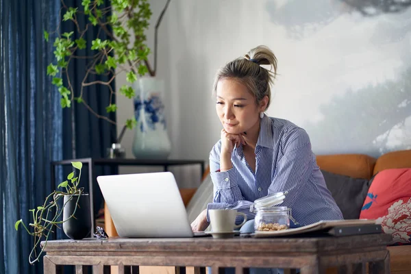 mature professional asian woman working from home sitting in couch looking at laptop computer