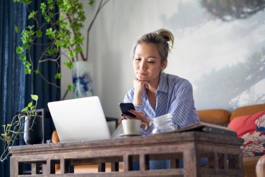 mature professional asian woman sitting in couch working from home using mobile phone and notebook computer