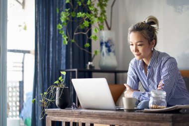 mature professional asian woman working from home sitting in couch looking at laptop computer