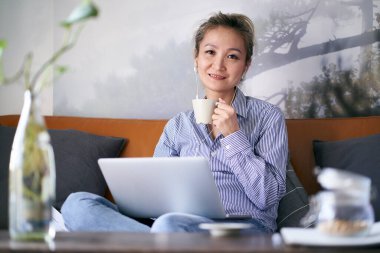 Yetişkin, profesyonel Asyalı bir kadın kanepesinde oturmuş elinde bir fincan kahveyle kameraya bakıyor.