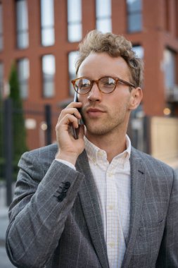 Yakışıklı, düşünceli, gözlüklü bir iş adamı. Cep telefonuyla konuşuyor. Sokakta dikiliyor, başka tarafa bakıyor.