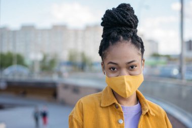 Siyah saçlı, koruyucu yüz maskesi takan ve sokakta kameraya bakan genç Afro-Amerikan kadın portresi. Sağlık hizmeti kavramı 