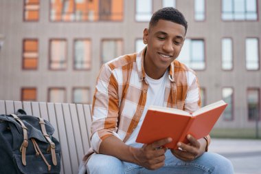 Kitap okuyan yakışıklı bir gencin portresi, parkta dinleniyor. Güler yüzlü Afro-Amerikan öğrenci üniversite kampüsünde oturuyor, eğitim kavramı öğreniyor.