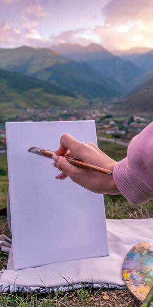 Photo A girl holds in her hand a paintbrush near a white canvas on the background of a mountain settlement on a summer evening. Scenic view, nature drawing concept.