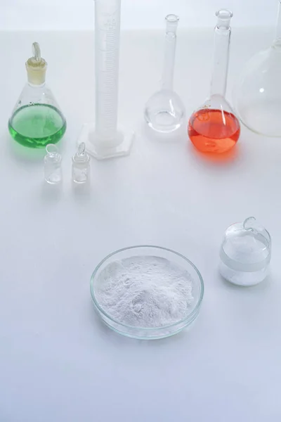 Boîte de Pétri avec poudre chimique blanche debout sur la table de laboratoire à côté de flacons isolés sur blanc — Photo