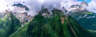 Panorama Caucasus mountains. Glacier turning into the river. Mountains in the clouds. Beautiful snow-capped mountains in summer.