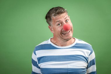 Kırmızı palyaço burunlu, gri saçlı, mavi-beyaz çizgili adam stüdyoda yeşil arka planda duruyor.