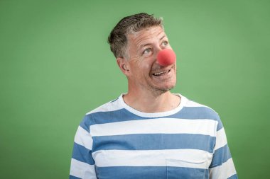 Kırmızı palyaço burunlu, gri saçlı, mavi-beyaz çizgili adam stüdyoda yeşil arka planda duruyor.