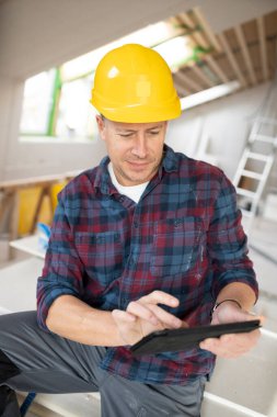 Sarı miğferli ve kareli gömlekli orta yaşlı bir adam tabletiyle çalışıyor.