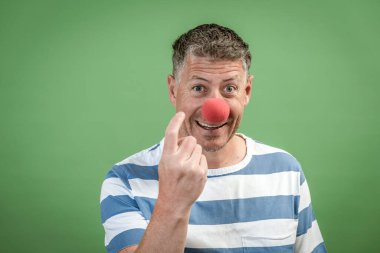 Kırmızı palyaço burunlu, gri saçlı, mavi-beyaz çizgili adam stüdyoda yeşil arka planda duruyor.