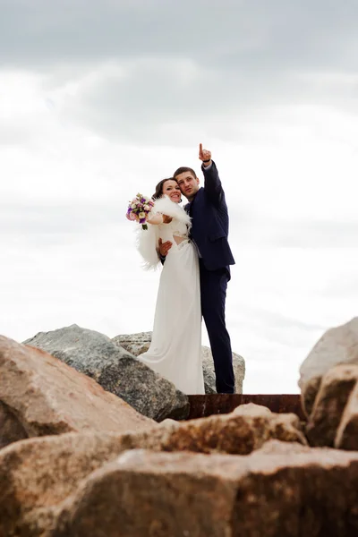 Happy newlyweds standing among the rocks on the beach. — Stock Photo, Image
