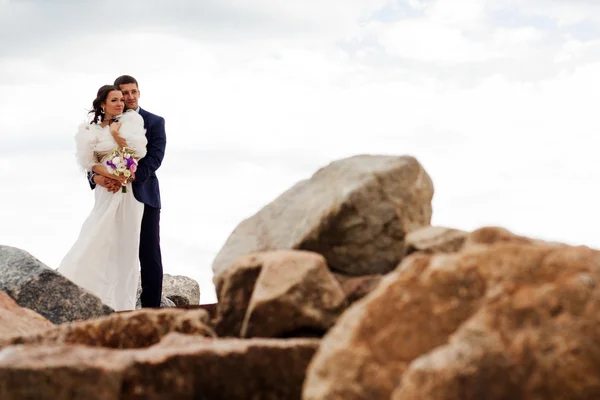 Happy newlyweds standing among the rocks on the beach. — Stock Photo, Image