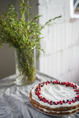 Masanın üzerinde beyaz mascarpone kreması ve kızılcıklarla süslenmiş lezzetli bir havuçlu kek duruyor. Her gün için güzel bir tatlı. Masadaki vazoda yeşiller var. Bahar hala hayat