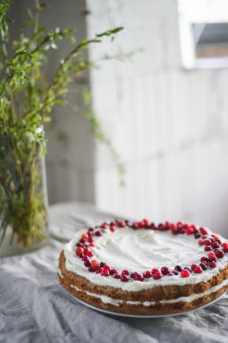 Masanın üzerinde beyaz mascarpone kreması ve kızılcıklarla süslenmiş lezzetli bir havuçlu kek duruyor. Her gün için güzel bir tatlı. Masadaki vazoda yeşiller var. Bahar hala hayat