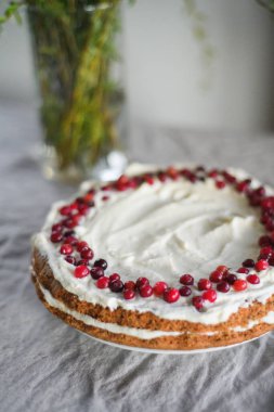 Masanın üzerinde beyaz mascarpone kreması ve kızılcıklarla süslenmiş lezzetli bir havuçlu kek duruyor. Her gün için güzel bir tatlı. Masadaki vazoda yeşiller var. Bahar hala hayat