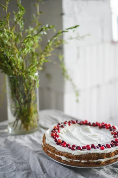 A delicious carrot cake stands on the table, garnished with white mascarpone cream and cranberries. A beautiful dessert for every day. There are greens in a vase on the table. Spring still life — Stock Photo, Image