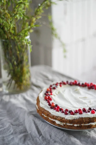 Kue wortel yang lezat berdiri di atas meja, dihias dengan krim mascarpone putih dan cranberry. Makanan penutup yang indah untuk setiap hari. Ada sayuran dalam vas di atas meja. Spring masih hidup — Stok Foto