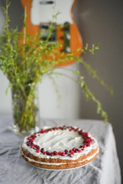 A delicious carrot cake stands on the table, garnished with white mascarpone cream and cranberries. A beautiful dessert for every day. There are greens in a vase on the table. Spring still life — Stock Photo, Image