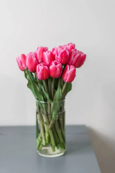 A large bouquet of pink tulips in a transparent vase stands on a blue table. A beautiful bouquet for a girl or for a holiday. Springtime abundance of flowers. Freshness and purity.