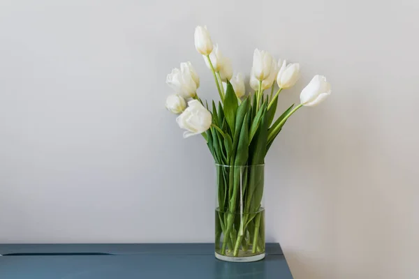 蓝色的桌子上立着一束漂亮的白色郁金香 插在一个透明的花瓶里 后面的轻墙和文字的地方 家庭内部 高质量的照片 — 图库照片