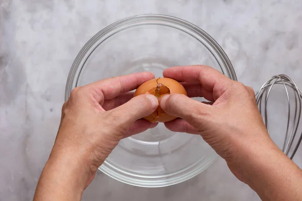 Вид зверху жінки рука розтріскує сире коричневе яйце в скляну миску на мармуровій поверхні — стокове фото